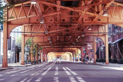 A view down the middle of Franklin Street, entirely empty of cars. The street is covered by the industrial elevated tracks of the “L” train. Patterns of light dapple on the empty gray street, creating an interplay of light and shadow. A single pedestrian crosses the expansive street.