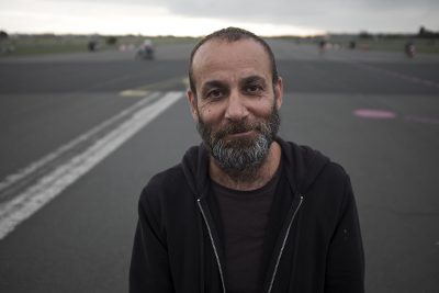 A medium skinned man with a beard stands in a road and smiles warmly into the camera.