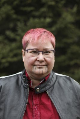 Headshot of TJ Cuthand. TJ is wearing a red top with a dark gray jacket. TJ is wearing glasses and has pink hair with a tattoo on his chin.